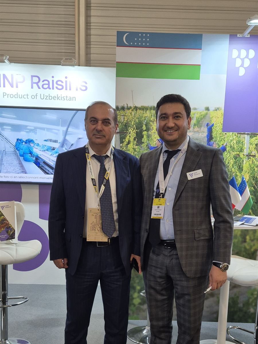 Two of the NNP Raisins team at the stand at SIAL Paris 2024, in front of a Product of Uzbekistan panel and the Uzbek flag, with the sorting-line footage on the screen beside them.